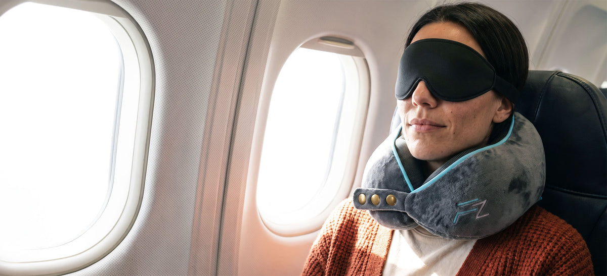 A woman wearing a black sleep mask and a gray neck pillow with blue trim sits in an airplane seat next to a window.